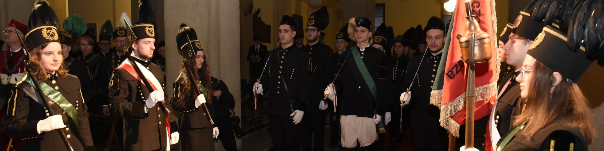 Zdjęcie z Uroczystości Barbórkowych w holu A0 AGH. Na zdjęciu studenci w mundurach górniczych. Po prawej  poczet sztandarowy ze sztandarem AGH. Naprzeciwko niego asysta z toporami. W śrdku Lis Major z asystą ze szpadami górniczymi.
